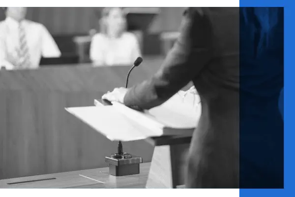 Black and white image of a woman standing in a courtroom at a lectern in front of microphone